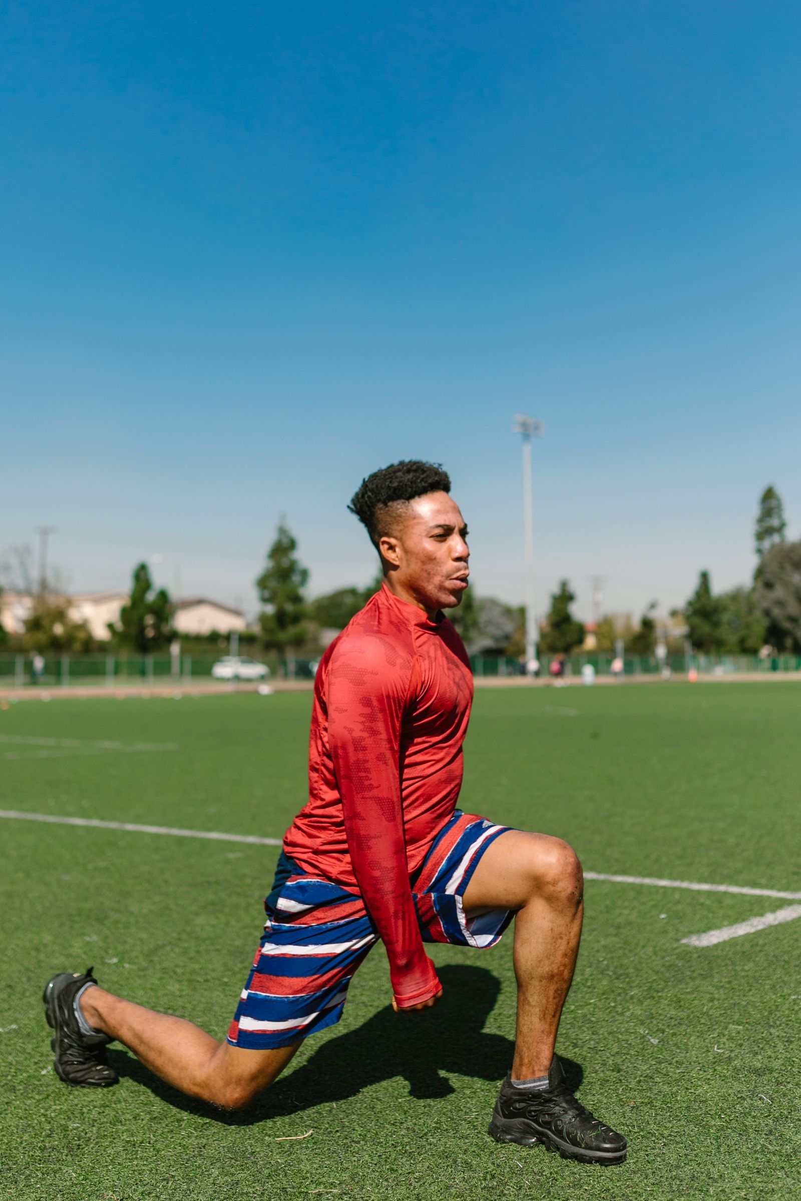 Runner in a red top doing a lunge warm-up on a sports field, the kind of activity that can trigger high hamstring tendinopathy and sit bone pain in Hinsdale, IL